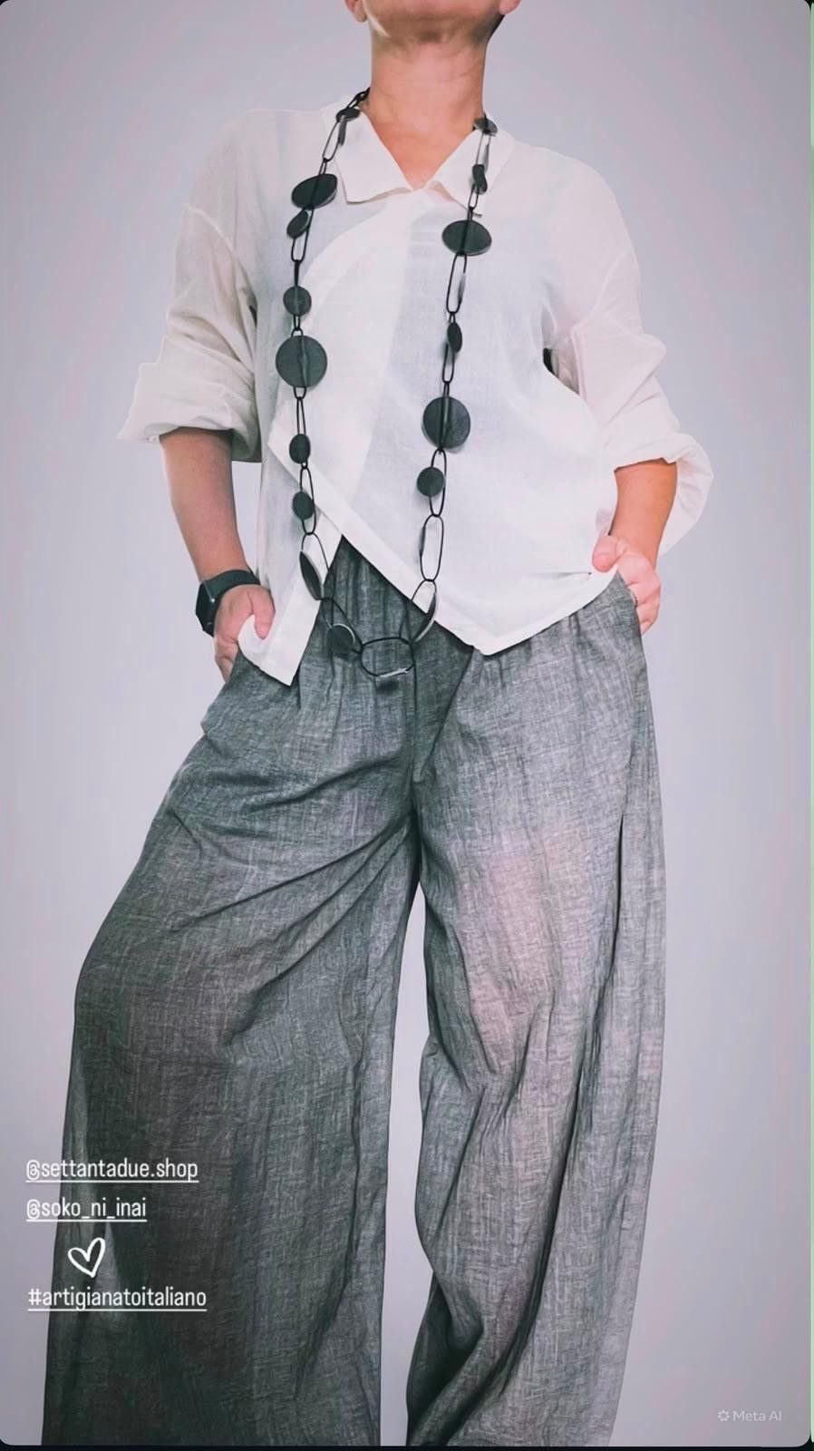 Camicia bianca leggera dal taglio asimmetrico, pantaloni ampi in lino grigio e collana lunga con dischi neri.