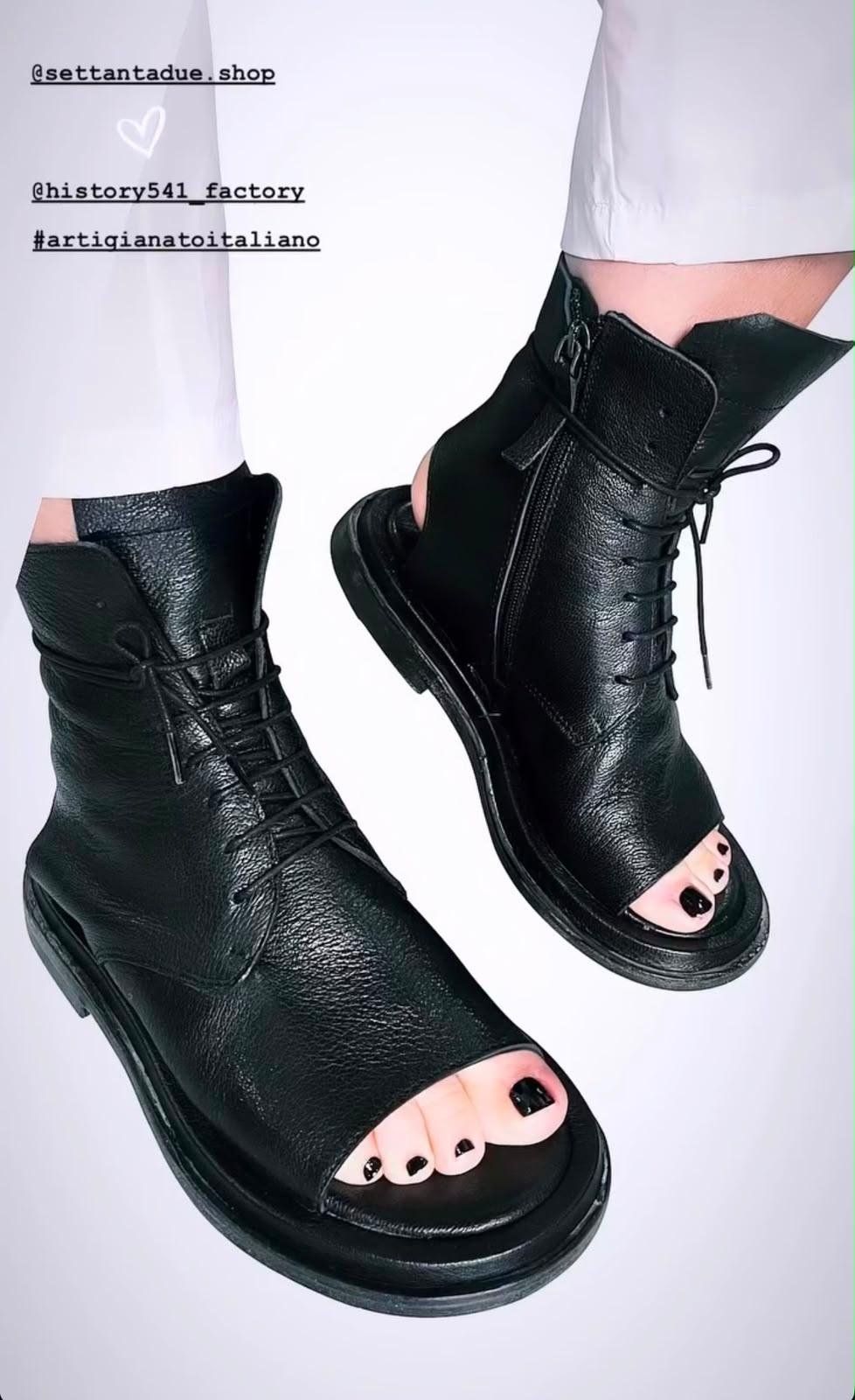 Anfibi in pelle nera con punta aperta, chiusura a lacci e zip laterale, indossati con un pantalone bianco.